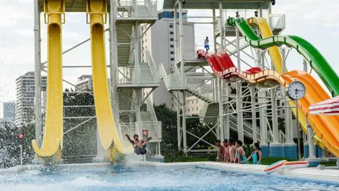A wide shot of water slides at a water park. A man is sliding off a large yellow slide while people watch in a line beside the pool.