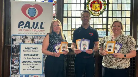 Three people holding a purple and a yellow leaflet stood in front of a stained glass window inside a building. They are stood next to a banner with multiple images and reads 'P.A.U.L For Brain Recovery'. The woman on the left is wearing a black spotty blouse and black trousers. She has long blonde hair. The man is wearing a black polo shirt with a red and blue logo on and black trousers. He has short brown hair. The woman on the right is wearing a black and white patterned blouse and black trousers. She has light-coloured hair tied back. They are all smiling at the camera. 