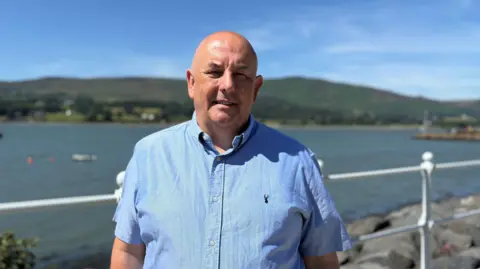 Councillor Jarlath Tinnelly standing on Warrenpoint promenade on a sunny day. He is bald and is wearing a light-blue short-sleeved shirt. The mouth of a calm, blue Carlingford Lough can be seen through white railings behind him. There are green hills in the distance and the edge of Warrenpoint Port is out of focus in the right-hand side of the photo.