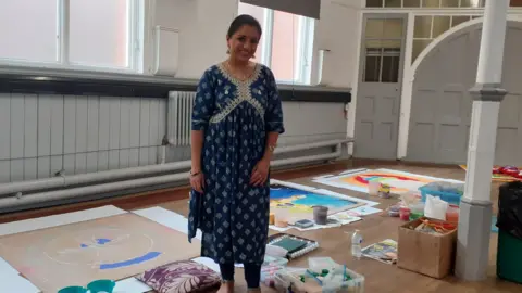 BBC A woman smiling in front of large artworks being created inside a community centre hall