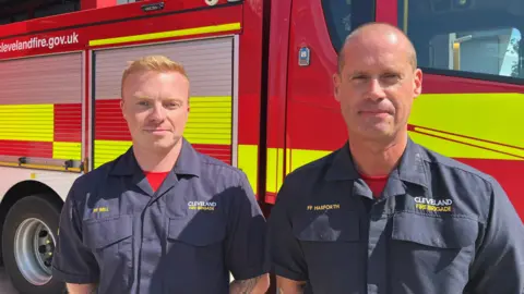 Two firefighters in uniform, in front of fire engine. 