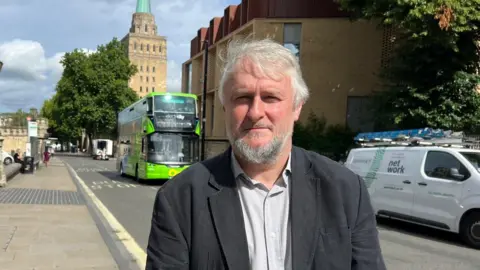 LDRS Councillor Epps stands beside a main road near a passing van and bus. He has light grey hair and a beard and wears an open-necked shirt under a dark jacket.