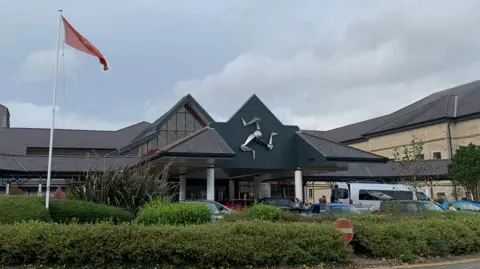 The entrance to Noble's Hospital, with the silver triskelion emblem on the front of the building and a Manx flag in front to the left above a green area of bushes and shrubbery.
