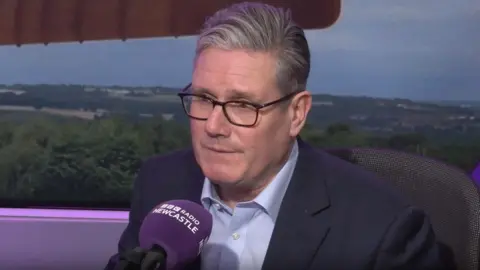 Prime Minister Keir Starmer sitting in front of a purple microphone in the BBC Newcastle studio. He has grey hair swept to the side and is wearing a suit and glasses.