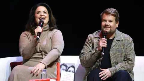 Getty Images Ruth Jones and James Corden sit on stage during the "When Gavin Met Stacey And Everything In Between" book event at the London Palladium. Ruth Jones talks into a black microphone. She is smiling and wears a cream coloured dress. She has shoulder length brown hair and is wearing gold hoop earrings and a gold necklace. James Corden also holds a microphone and is wearing a checked blazer with a black t-shirt and black trousers. Their book sits on a table in front of them.