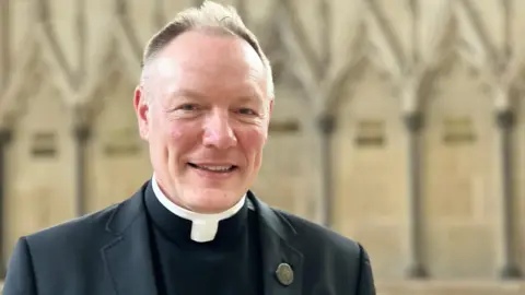 The Very Rev Toby Wright. He has short grey hair and no facial hair. He is wearing black and white clerical clothing and is looking at the camera and smiling.