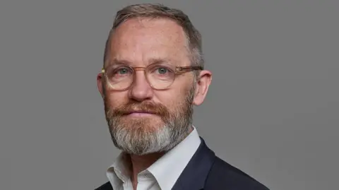 James Purnell, un hombre de mediana edad con barba pelirroja y canosa y gafas, posa para una fotografía formal.