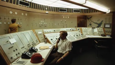 Getty Images Operators at work in the control room of the Cruachan hydro-electric scheme in 1974. One of the operators is holding phone to an ear as he sits behind a bank of controls covered in switches and dials.