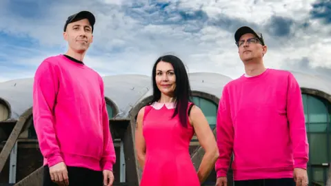 Bis The band Bis - Steven Clark, Amanda McKinnon and John Clark - all dressed in pink and looking directly towards the camera