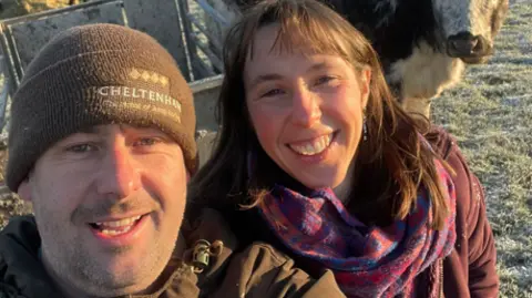 A man and woman stand next to each other in a frost covered field, and look up smiling to the camera. There is a cow behind them