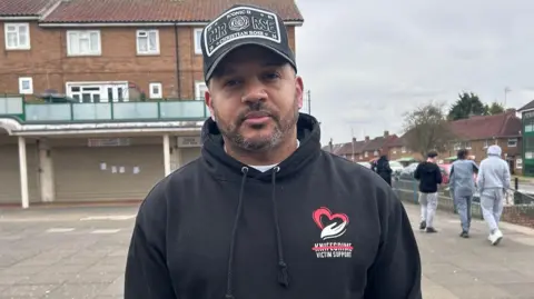 Kate Bradbrook/BBC Quinton Green looks at the camera wearing a black hoodie with a logo showing a red heart turning into a white flame. The work KNIFECRIME is ruled out and the words Victim Support below. He has a black baseball cap and a neat beard and moustache. Behind him some young people walk past. He is at a 1980s style concrete shopping precinct with housing/flats above.