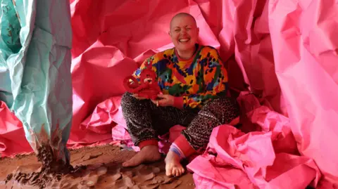 Sculptor Lady Kitt. She is wearing a colourful jumper and animal print trousers. She has a shaved head and is smiling widely at the camera. She is sitting on a piece of large pink fabric which runs from the floor vertically up behind her. Her bare feet are resting on ground made of brown clay.