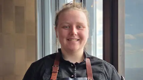 Holly smiles at the camera and has her blonde hair tied in a bun. She wears a black shirt and a brown apron. 