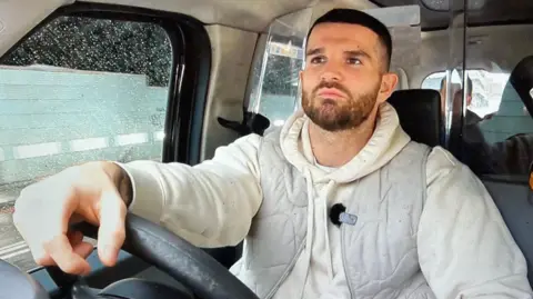 Tom Williams/BBC Jack Cawley is wearing a grey bodywarmer over a cream hoodie. He has short black hair and a beard. He is at the wheel of his black cab.