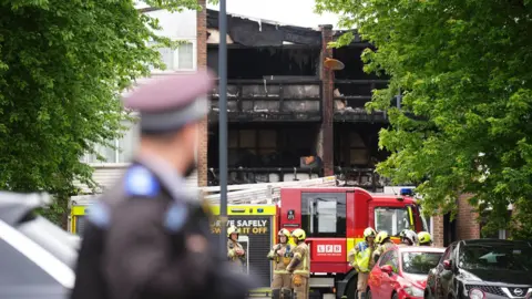 PA Media a police officer looks back at the two houses devastated by the fire, as fire crews and a fire engine wait outside