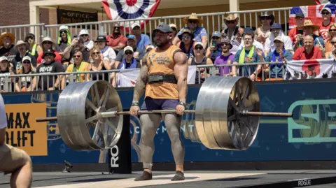 Shane Flowers came 7th in the World's Strongest Man contest in California. Here he is at the contest deadlifting a bar with a large amount of weight on. crowds watch on.