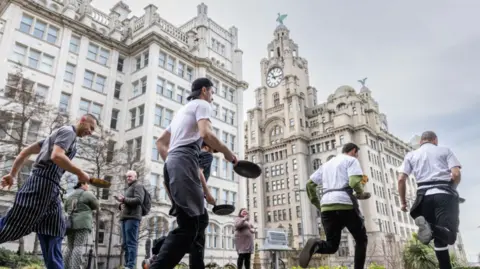 Shot of several chefs in their chef whites running round the church gardens with pancake and pan in hand. The Liver Building can be seen in the distance.