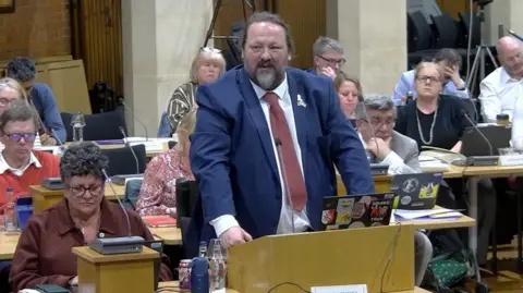A councillor stands at a wooden lectern speaking in a council chamber. Rows of desks with seated councillors, papers, laptops and bottles of water are visible behind, with the meeting taking place in a large indoor hall.