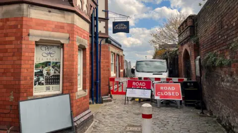 A red brick building on the left with a black hanging sign that reads "Cliff Railway". In the walkway next to it, are red and white barriers preventing people from walking. There are red signs that say "road closed"