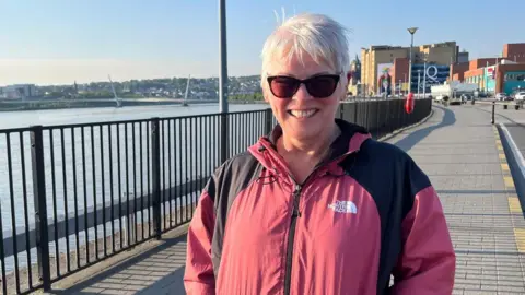 Pauline is smiling at the camera. It is a very sunny day, and she is wearing brown sunglasses. She has short white hair and is wearing a pink 'North Face' coat.