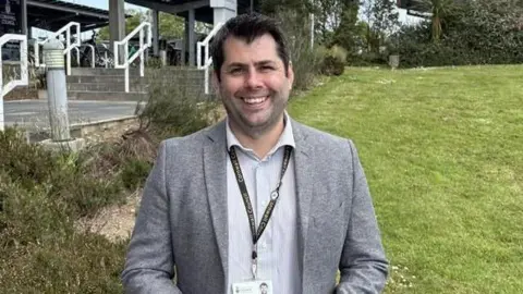 Leigh Frost weaning a grey jacket and shirt smiling in front of the Cornwall Council building