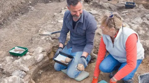 Teffont Archaeology Two archaeologists on the ground in a dig with the foundations of old walls around them. The walls look like pieces of old plaster with bits of colour on.