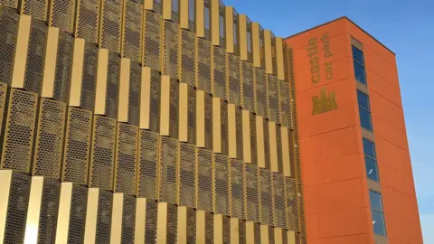 A multi-storey car park, with gold panelling and a terracota-coloured cladded stairwell to the right. There is a gold image of a castle underneath lettering which reads Castle Car Park.