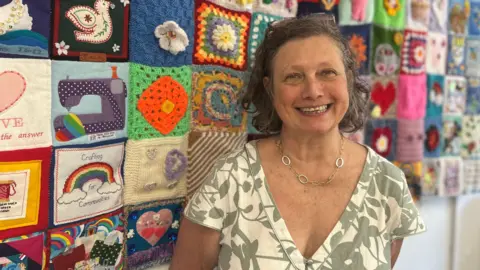 A woman in a v-neck white and green dress stands in front of a colourful crocheted blanket hung on a wall, smiling. She has bobbed curly brown hair, brown glasses pushed back on her head, and a silver chain link necklace.