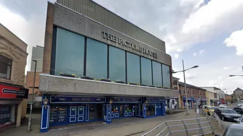 Google Exterior view of The Picture House building on Bethcar Street in Ebbw Vale. It has a wide path outside the entrance. It has a grey concrete facade, large upper windows and a blue-painted pub entrance.