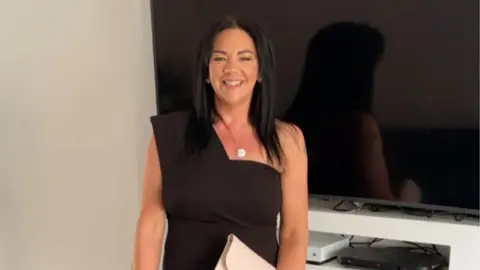 A woman standing in front of a TV and white wall, she is wearing a black dress and has black hair