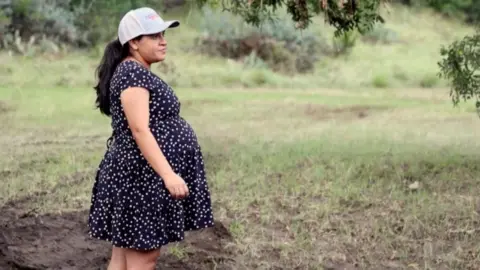 Heavily pregnant woman stands wearing a polka dot dress and a cap 