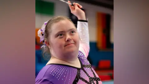Contributed A young woman pictured as she strikes a gymnastic pose. She holds a stick with a ribbon attached high above and behind her. She is wearing a pink and black leotard. 