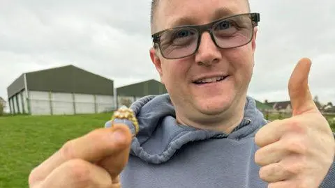 A man in his early 40s smiles as he stands in a field on a cloudy day. In one hand he is holding up a gold diamond ring, while he gives a thumbs up with the other hand. He is wearing a grey hoodie and black-rimmed glasses, the lens of which are slightly tinted. There are large metal barns behind him.