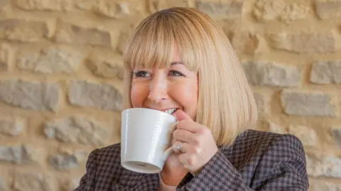 Cotswold Marketing Agency Michelle Niziol, who is wearing a chequered blazer and has her hair in a tight blonde bob, grins while holding a white mug up to her face.
