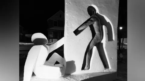 A black and white photo of a snow sculpture depicts a wall with a 2D cut out of a person. Next to it is a 3D person holding their hand up to the 2D cut down. A man can be seen nude stood in the 2D cutout.