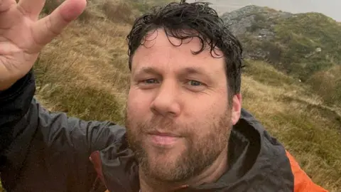 Selfie style picture of a bearded man with dark wavy hair on a hill. He is wearing a black and orange jacket and waving at the camera