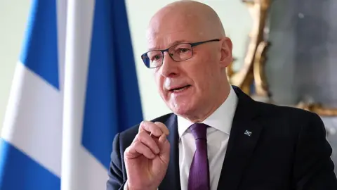 John Swinney, a blad man waring glasses, a dark suit with white shirt and purple tie, is standing in front of a Saltire flag with his right hand raised as he answers a question from an unseen group