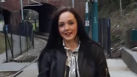 Lincolnshire Police Ellen Marshall with dark brown long hair and wearing a black bomber jacket over a monochrome striped dress. She is standing on a train platform with the train tracks visible on the left