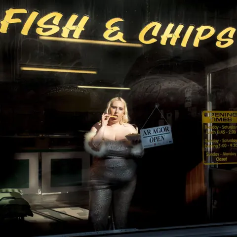 Megan Winstone Shot from outside a chip shop, Gina Tonic is wearing a silver strapless jumpsuit and is eating chips