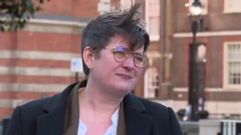 Olivia Bailey stands outside parliament for an interview, it's slightly blurred in the background. She has short mousy brown hair, spike at the front and wears round brown wire framed glasses, a black woolen coat with a brown blazer and white shirt underneath