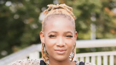 Contributed Ms Tanaka smiles at the camera. Her hair is tied back, she has a nose ring and wears an animal print top with a black pinafore top over it.