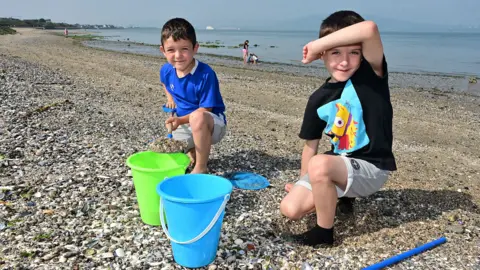 Pacemaker Two boys on a beach. They are both in shorts and T-shirts and have dark hair. There are two buckets in front of them - a green and blue one. A fishing net is also beside one of them.