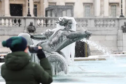 PA Media A man takes a photograph of an ice-covered mermaid statue in Trafalgar Square, London, on 9 February 2021