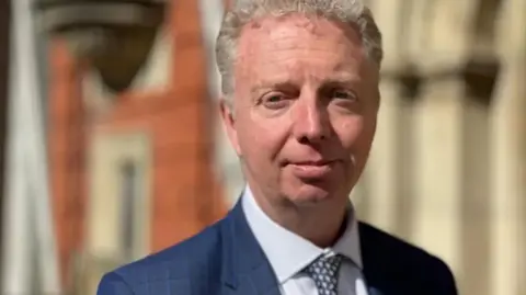 Alex Allinson, who is wearing a blue suit with a shirt and tie and has short grey/blond hair, outside the House of Keys.