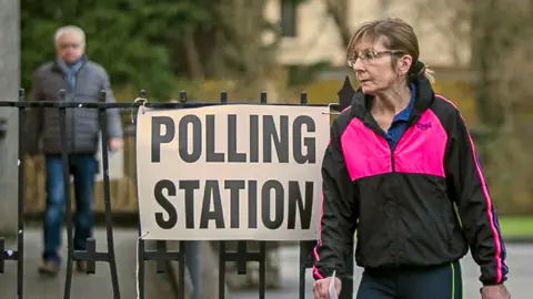 PA Media A woman stands by a sign for a polling station in Northern Ireland