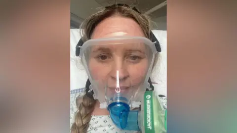 Supplied Aimee is sitting on a hospital bed and is taking a selfie. She has long blonde hair in braids and is wearing an oxygen mask which is covering her whole face.