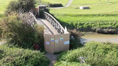Sussex News and Pictures A metal bridge over a river. The entrance to the bridge is blocked off by a wooden barrier.