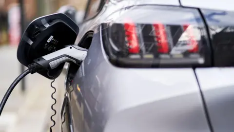 A silver car parked on the street, being charged. 