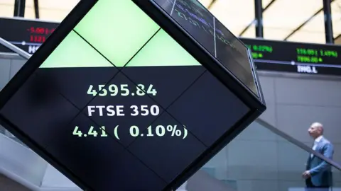 Getty Images a bald man in grey suit walks past a sign showing share prices in the London Stock Exchange