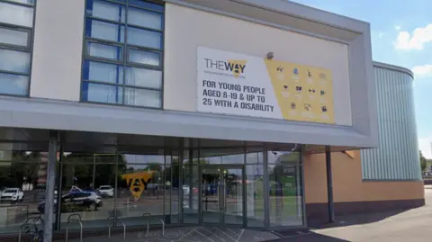 A large building with glass windows. It has a large yellow and white poster on the wall saying 'The Way' on it as well as details about the organisation.